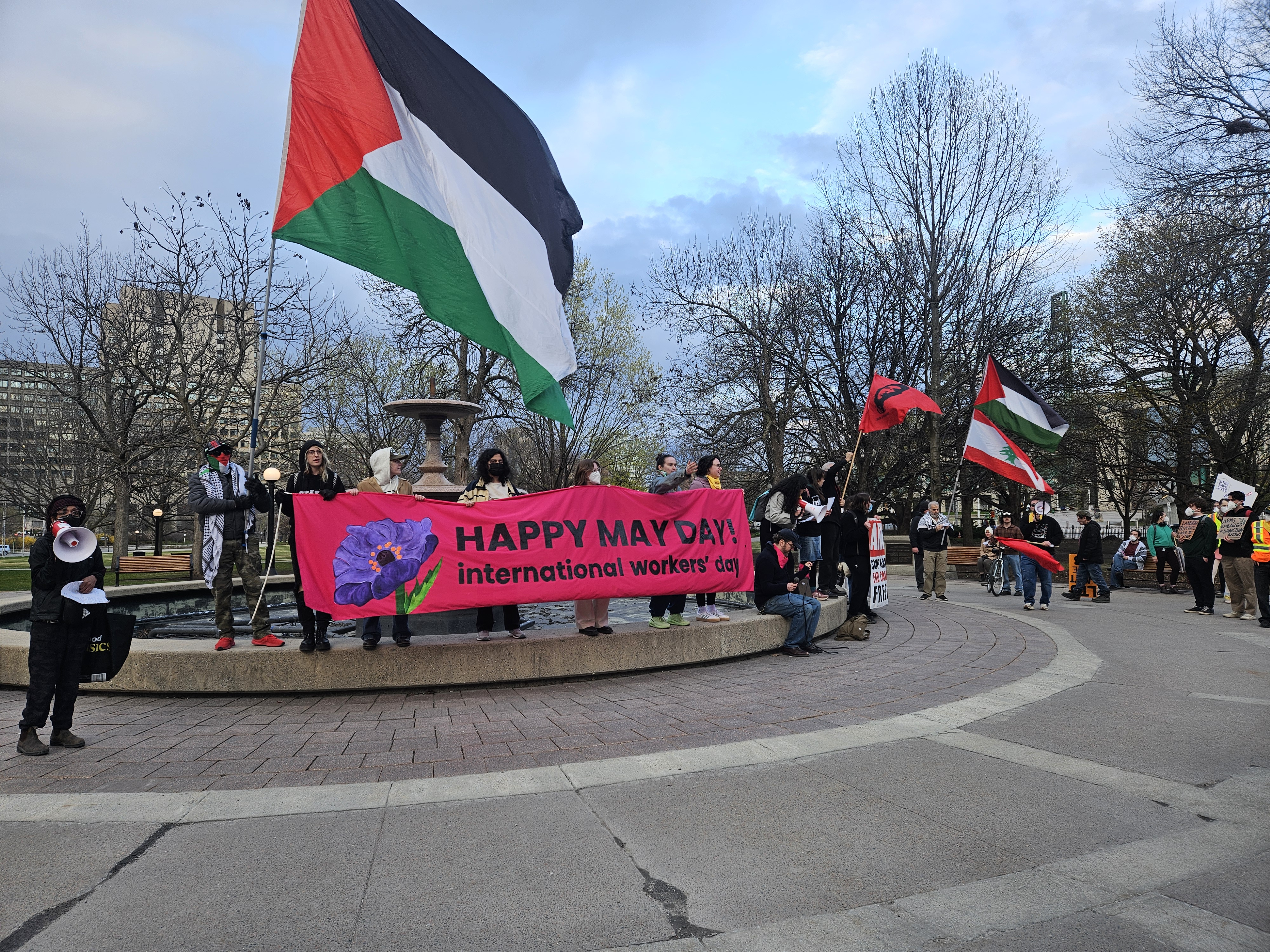 Some people holding a bright pink banner that says "Happy May Day! International Worker's Day" A few others holding Palestine flags, and a Lebanon flag.